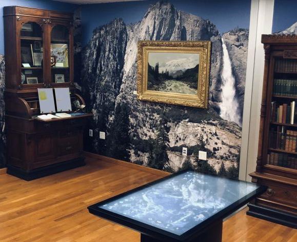 a room with an antique desk and bookcase filled with old books, a painting of the Sierra Nevadas, and a digital screen for viewing information